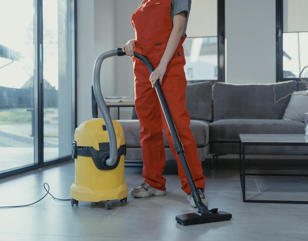 A professional cleaner in red coveralls vacuuming a modern living room with a yellow vacuum cleaner.
