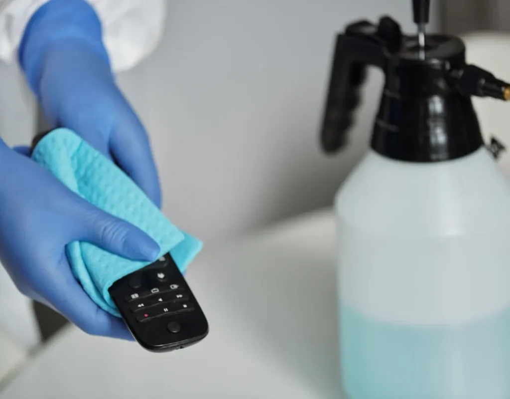 Gloved hands disinfecting a remote control with a cloth and spray bottle during medical office cleaning in Bentonville, AR. Medical Office Cleaning Near Me.